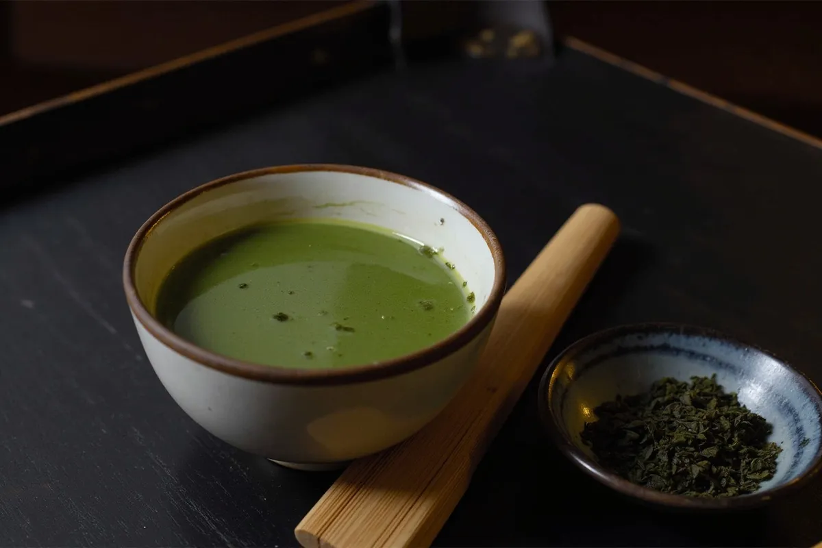 Matcha green tea in a ceramic bowl with a bamboo whisk beside a dish of loose tea leaves
