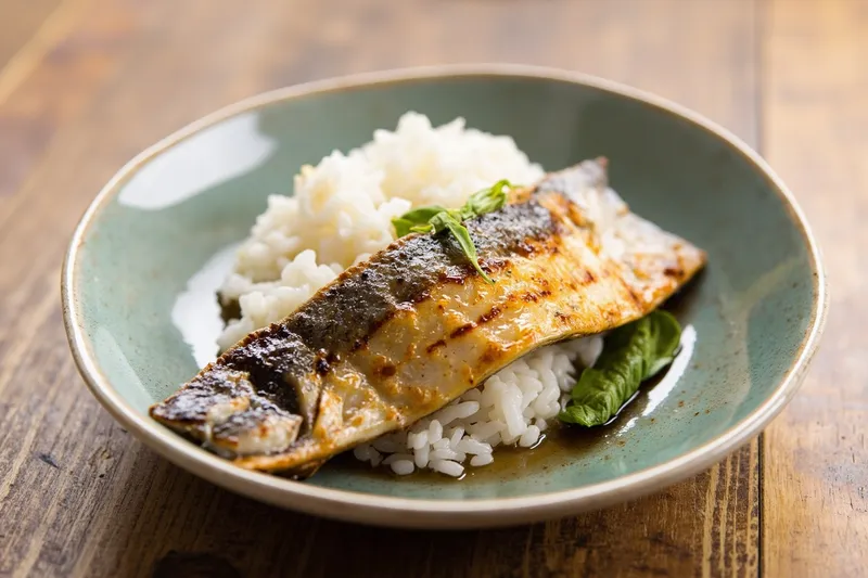 A white plate holding a browned grilled mackerel fillet next to a bowl of steamed white rice.