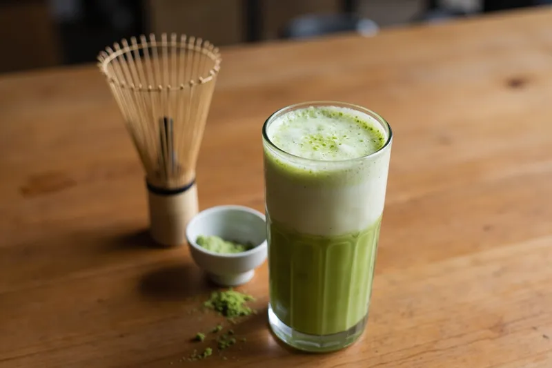 Matcha latte in a tall glass with a bamboo whisk and matcha powder on a wooden table