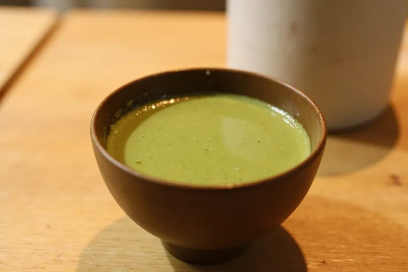 Green matcha in a ceramic bowl beside a bamboo whisk on a wooden table